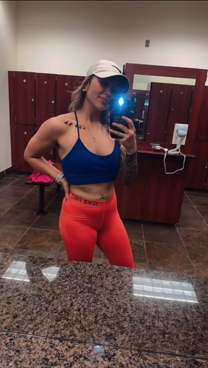 Fit woman in gym, taking selfie wearing sports bra and leggings. Locker room setting, holding phone.