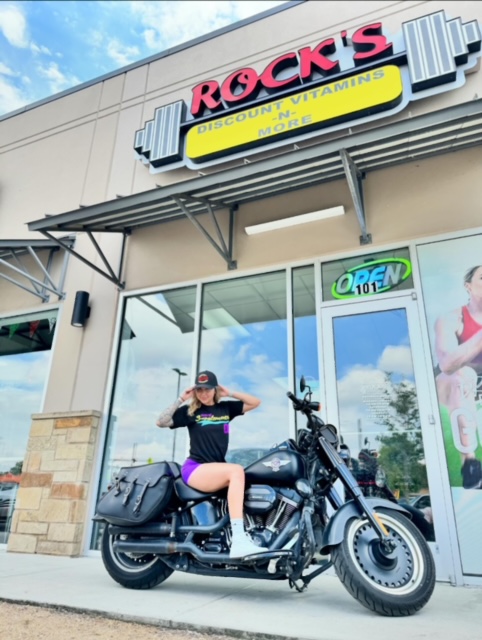 Alt text: Woman posing on motorcycle in front of Rock's discount vitamins storefront, open sign visible